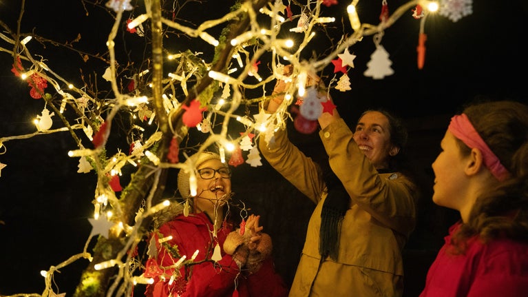 Woman and two children hanging baubles on a tree covered in fairy lights.